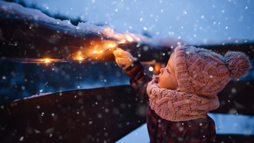 A toddler child in coat looking at lights on wooden railing at nighttime.